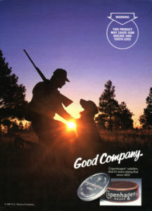 Copenhagen smokeless tobacco advertisement, featuring a silhouette of a man with a hunting rifle and a dog against a sunset or sunrise and trees "Good Company"